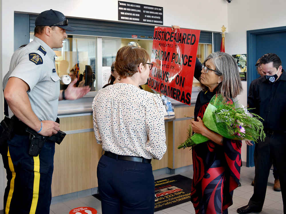 A woman with medium-light skin tone, grey hair and glasses holds a bouquet of flowers in one arm and in the other holds up a red sign with black lettering that reads, 'Justice 4 Jared Advocates to Disarm Police' and 'Always-On Body Cameras.' She is holding the sign towards a uniformed RCMP officer at the left of the frame.